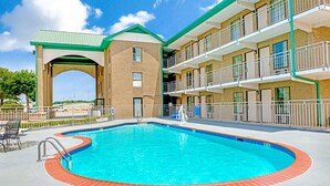 A swimming pool with a brick border, surrounded by a building with multiple balconies and a green roof.
