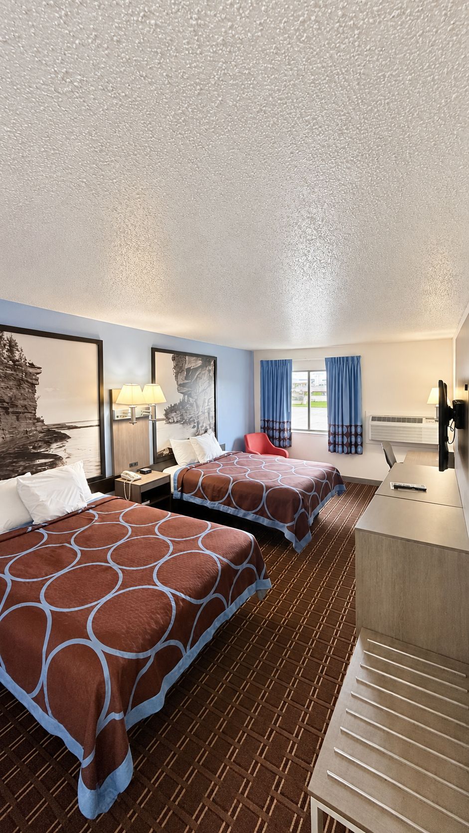 A hotel room with two beds, a desk, a chair, a lamp, and a window with curtains.