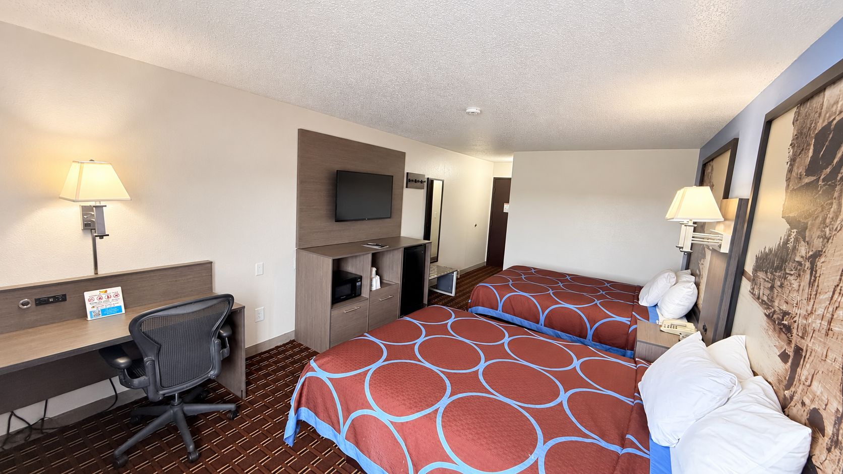 Quarto de hotel com duas camas, uma escrivaninha, uma cadeira, uma televisão e um quadro na parede.