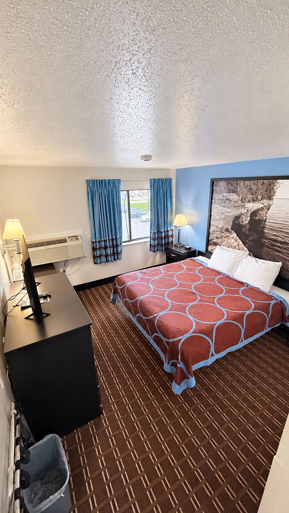 A hotel room with a bed, a desk, a chair, a lamp, a window with curtains, and a wall-mounted air conditioning unit.