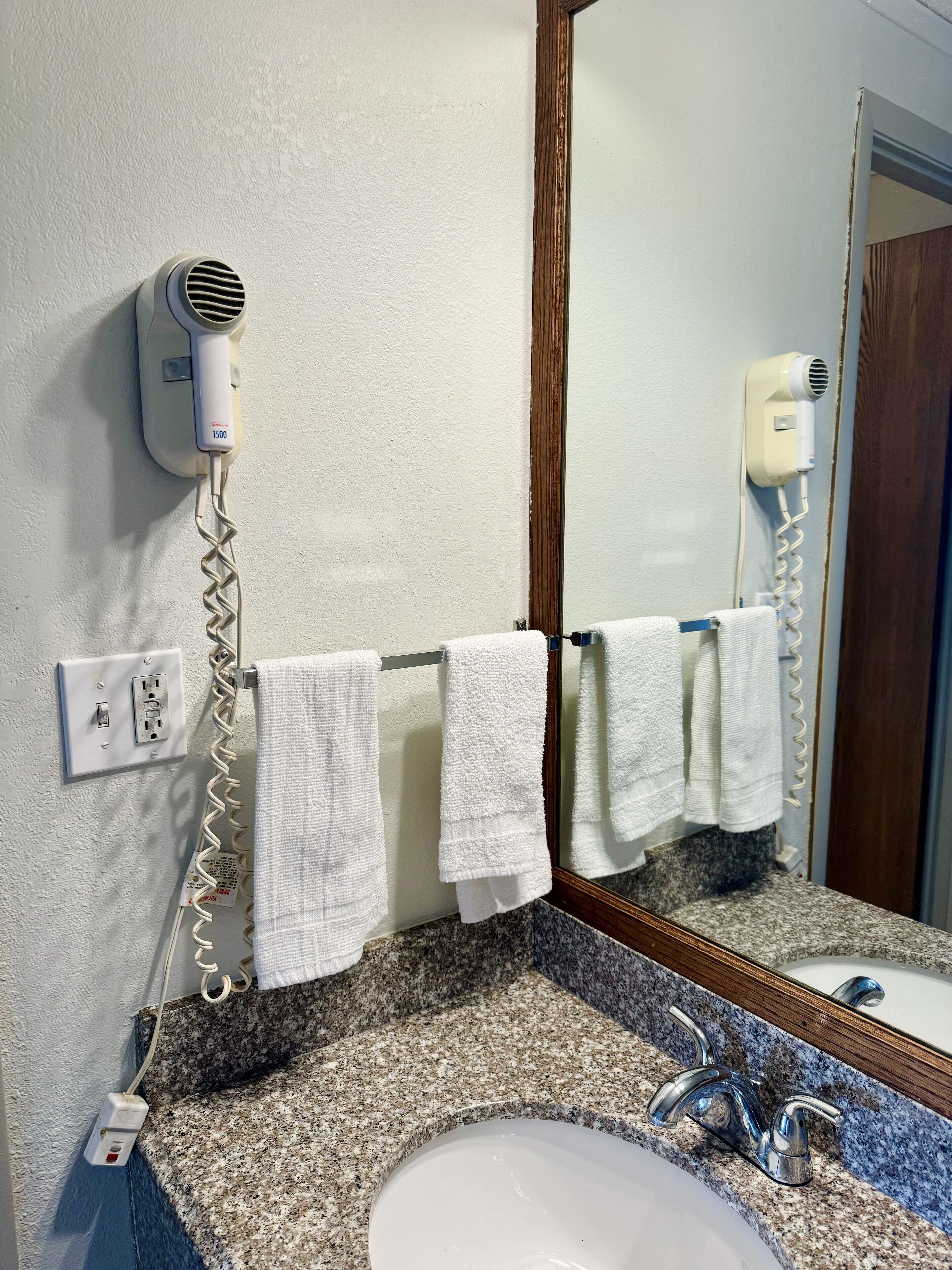 Ein Badezimmer mit Granitplatte, einem weißen Waschbecken, einem Spiegel und zwei an der Wand montierten Haartrocknern.