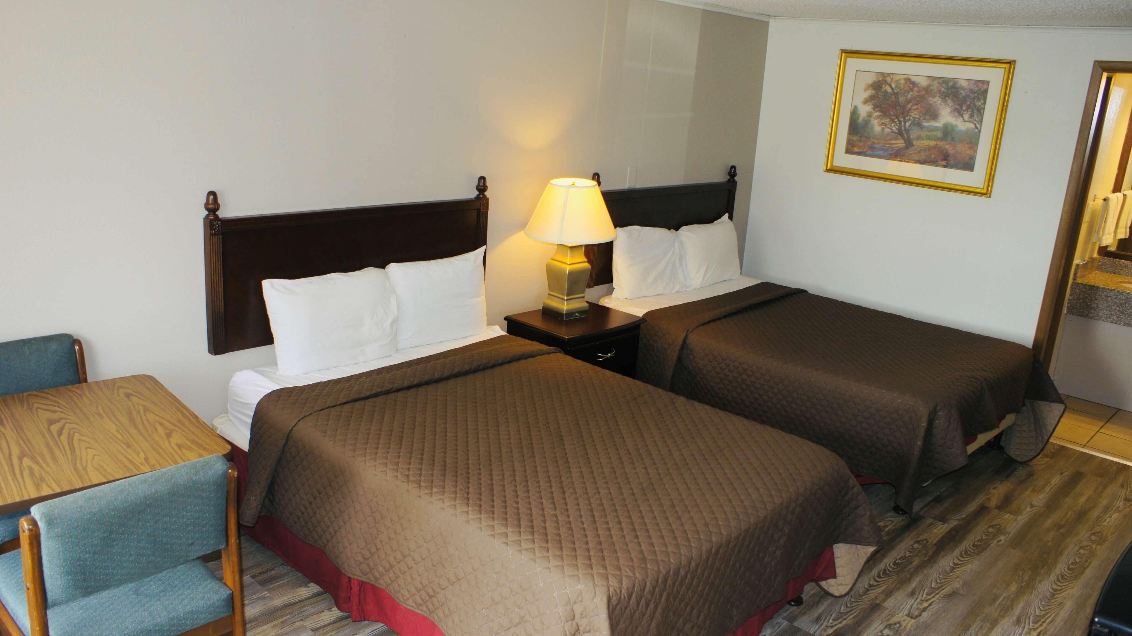 Ein Hotelzimmer mit zwei Betten, einem Nachttisch mit Lampe, einem Holzstuhl und einem Gemälde an der Wand.