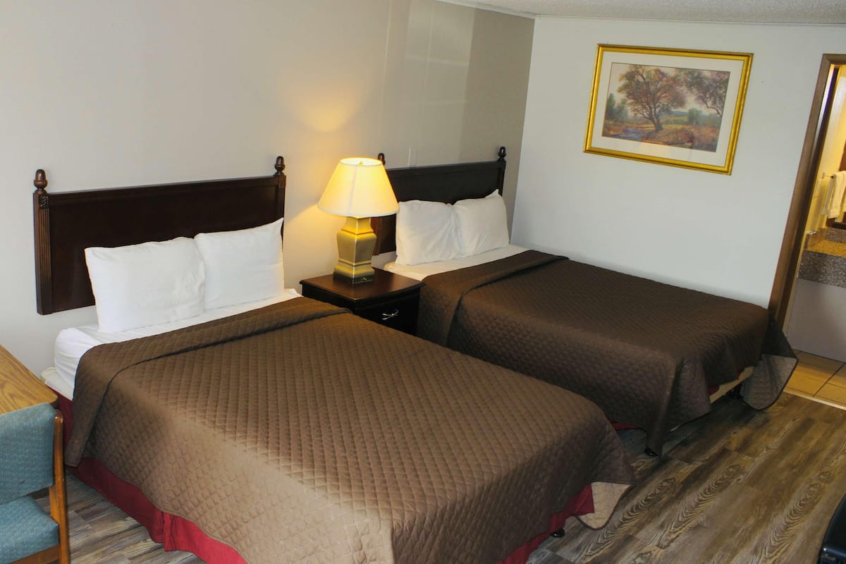 Camera d'albergo con due letti, un comodino con una lampada, una sedia in legno e un quadro appeso al muro.