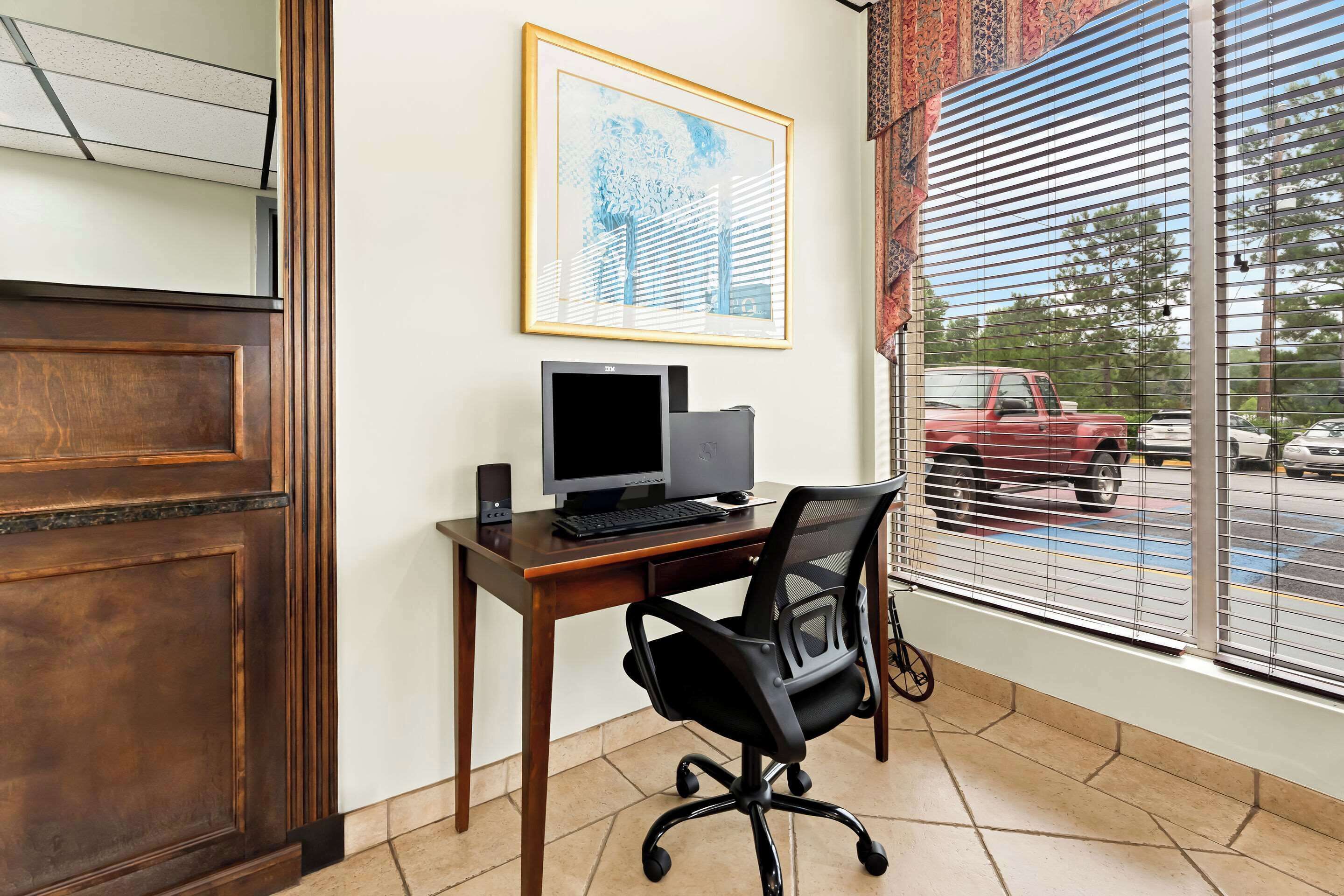 Une pièce avec un bureau en bois, un ordinateur et une chaise. Il y a une fenêtre avec des stores et une vue sur la rue.