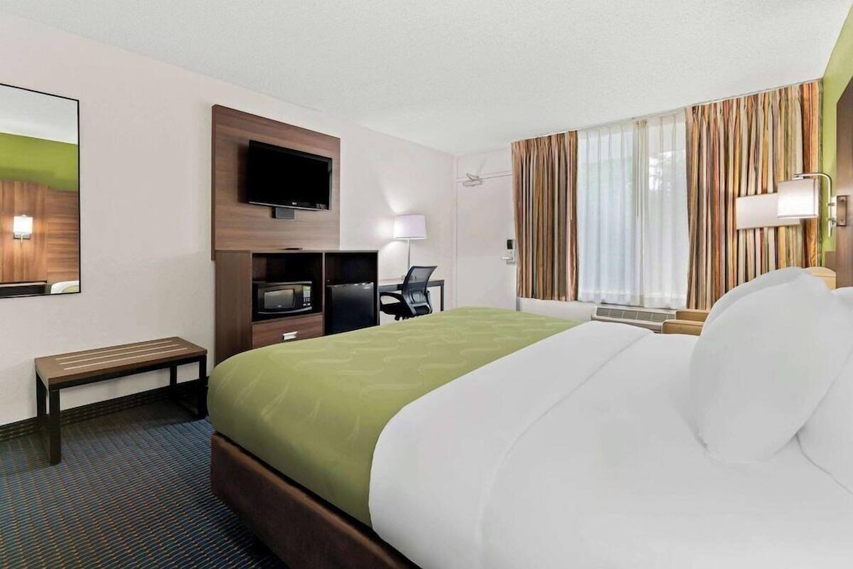 Quarto de hotel com uma cama grande, uma secretária com cadeira, televisão de tela plana, abajur e janela com cortinas.