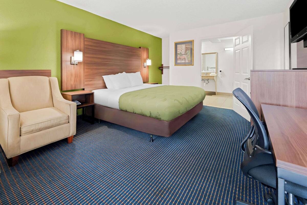 Quarto de hotel com cama, poltrona, escrivaninha e televisão.