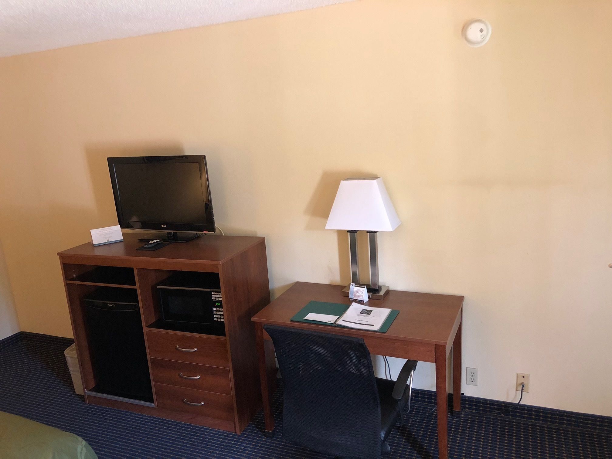 A hotel room with a wooden desk, a television, a lamp, a chair, and a trash bin.