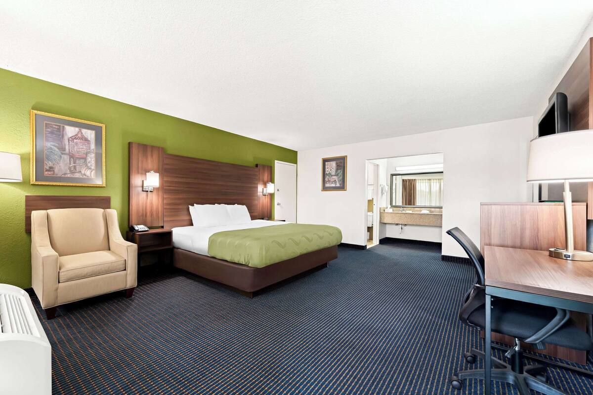 Quarto de hotel com cama, escrivaninha, cadeira e mesa de cabeceira.