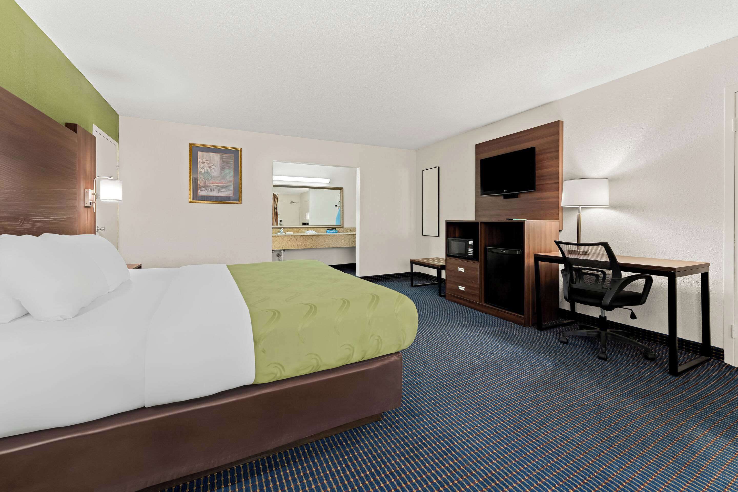 A hotel room with a bed, a desk, a chair, a television, and a bathroom visible in the background.