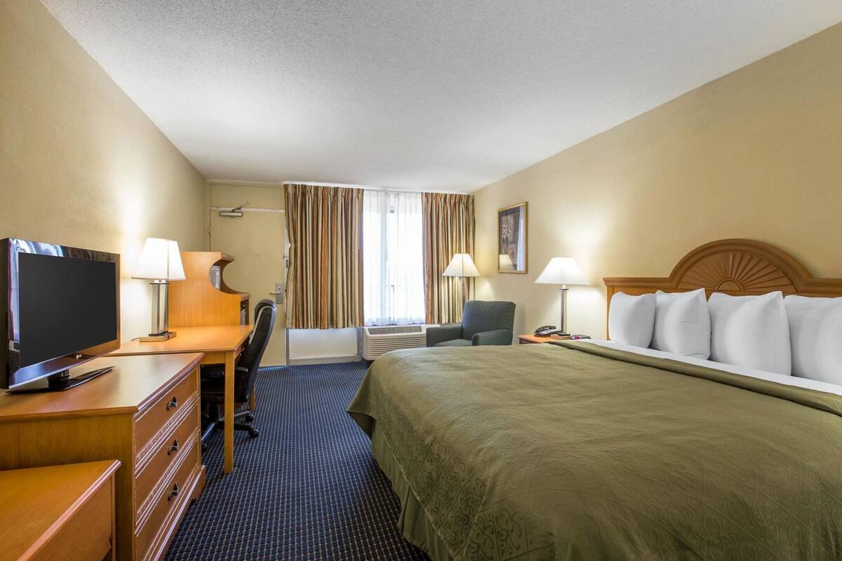 Quarto de hotel com cama, escrivaninha, cadeira, televisão, luminárias e janela com cortinas.