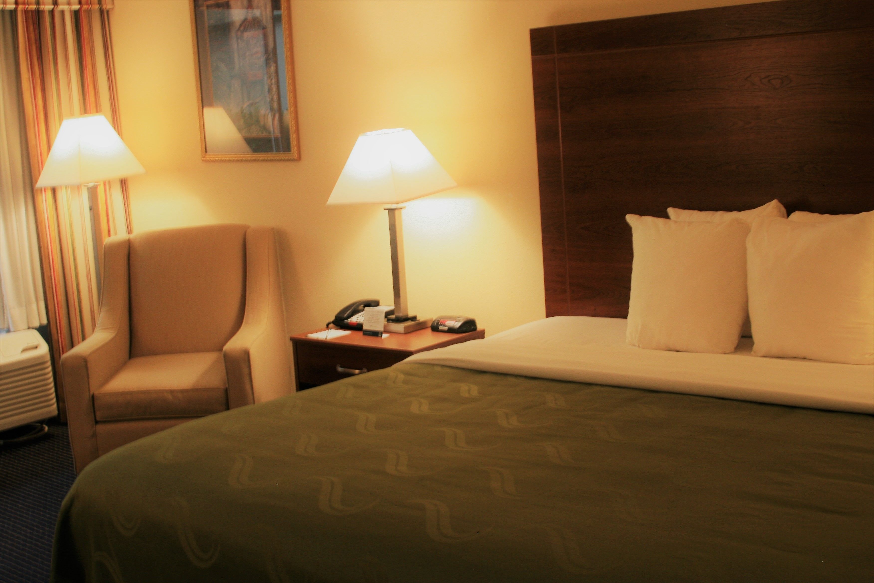 A hotel room with a bed, two bedside lamps, a chair, a nightstand, and a painting on the wall.