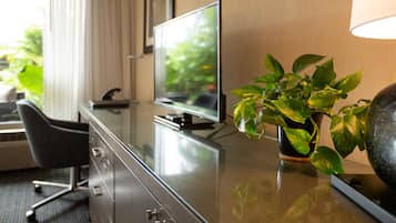 A hotel room with a flat-screen TV on a glass table, a potted plant, a desk with a chair, and a lamp.