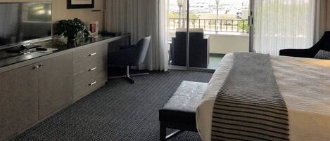 Ein Hotelzimmer mit Bett, Schreibtisch, Stuhl, Fernseher und Blick auf die Stadt durch große Fenster.