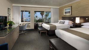 A hotel room with two beds, a desk, a chair, and a TV. There is a view of palm trees and a marina through the windows. - West Beach Inn, a Coast Hotel (Santa Barbara)