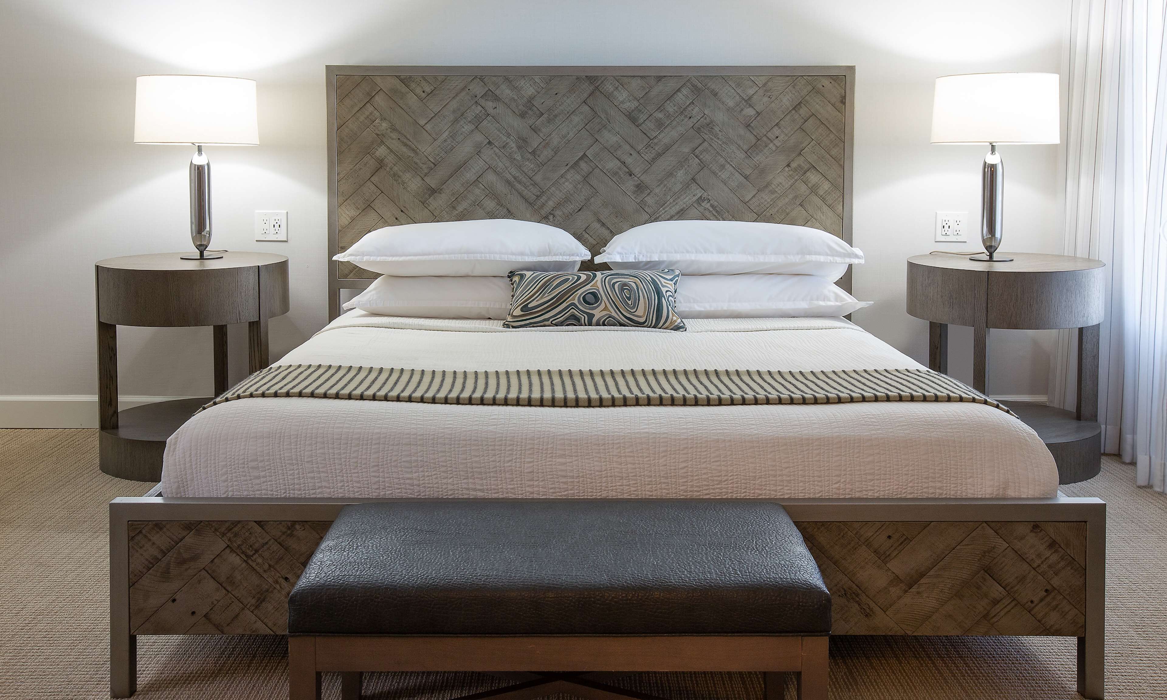 A neatly arranged hotel room with a large bed, two bedside tables with lamps, a bench at the foot of the bed, and a herringbone-patterned headboard.