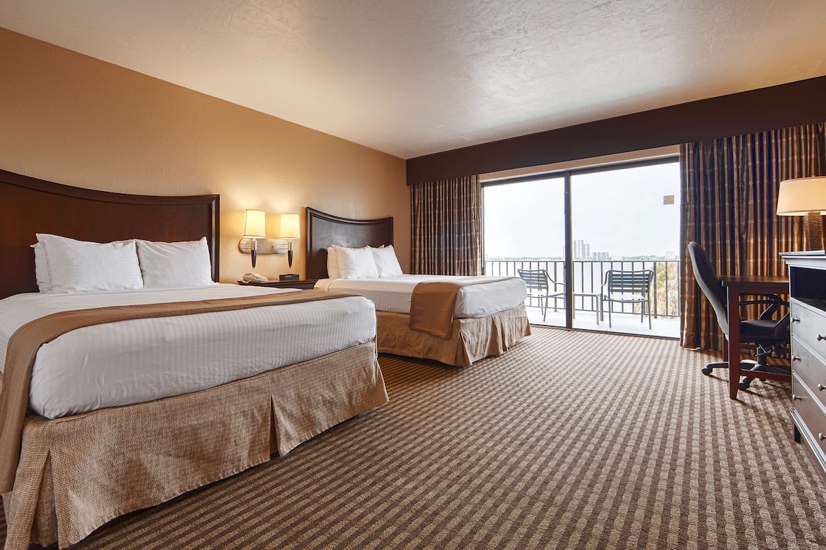 A hotel room with two beds, a large window with curtains, a balcony with chairs, and a desk with a lamp.