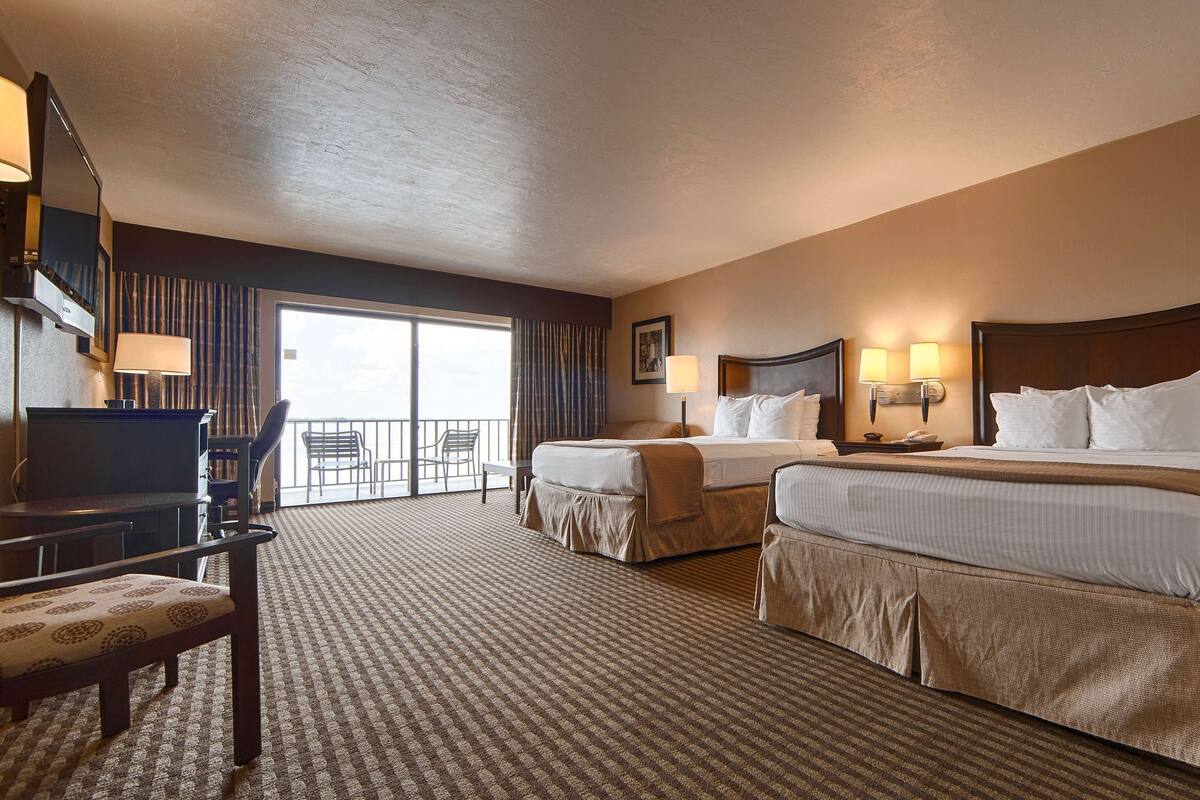 A hotel room with two beds, a desk, a chair, and a balcony with a view.