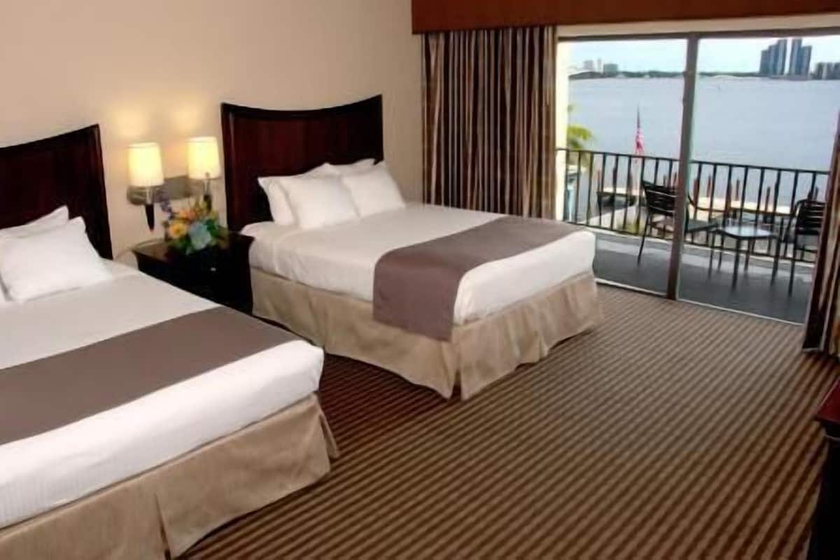 A hotel room with two beds, a nightstand with lamps, a view of the water and city skyline through the balcony.