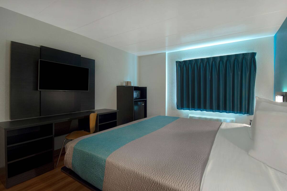 A modern hotel room with a large bed, a flat-screen TV mounted on the wall, a desk with a chair, and a window with curtains.