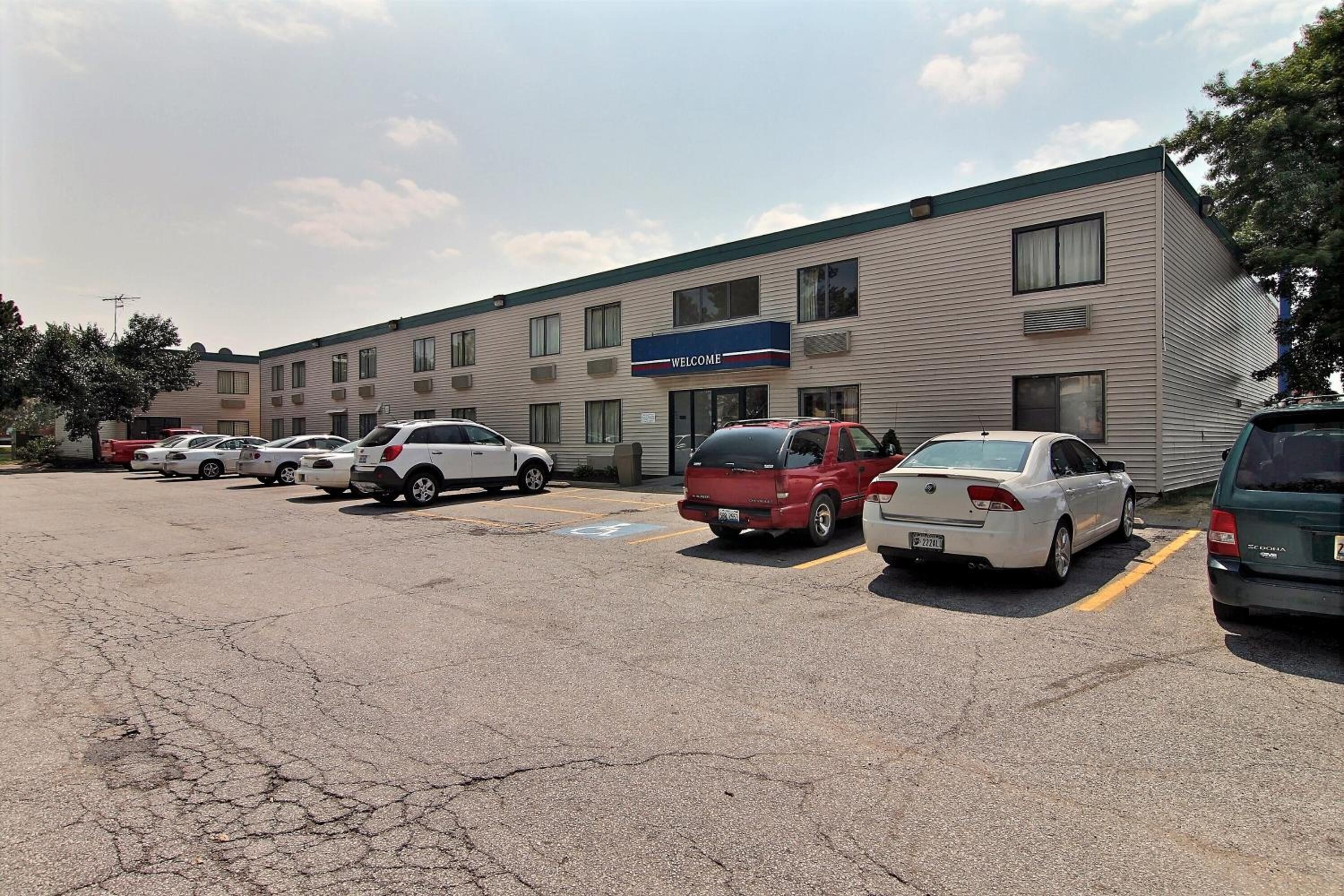 A multi-story building with a 'WELCOME' sign, parking spaces, and a row of parked cars.