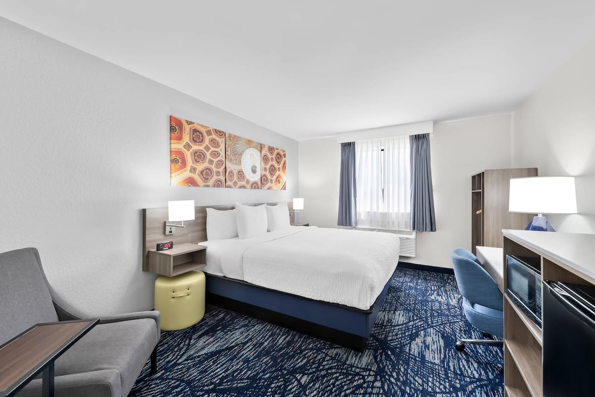 A hotel room with a bed, a desk, a chair, a small table, a TV, and a window with curtains.