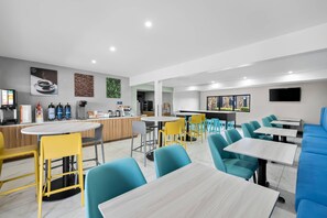A modern cafe with round tables, yellow and blue chairs, and a kitchen area with appliances.
