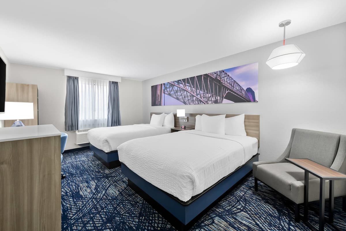 A hotel room with two beds, a desk, a chair, a large window with curtains, a wall-mounted TV, and a painting of a bridge.