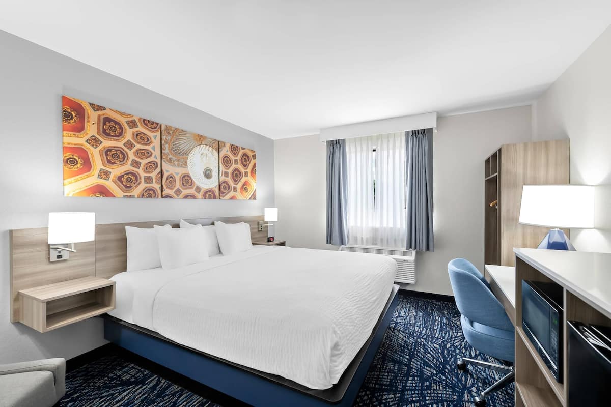 A hotel room with a large bed, a desk with a computer, a chair, a wardrobe, a window with curtains, and a wall with a patterned artwork.