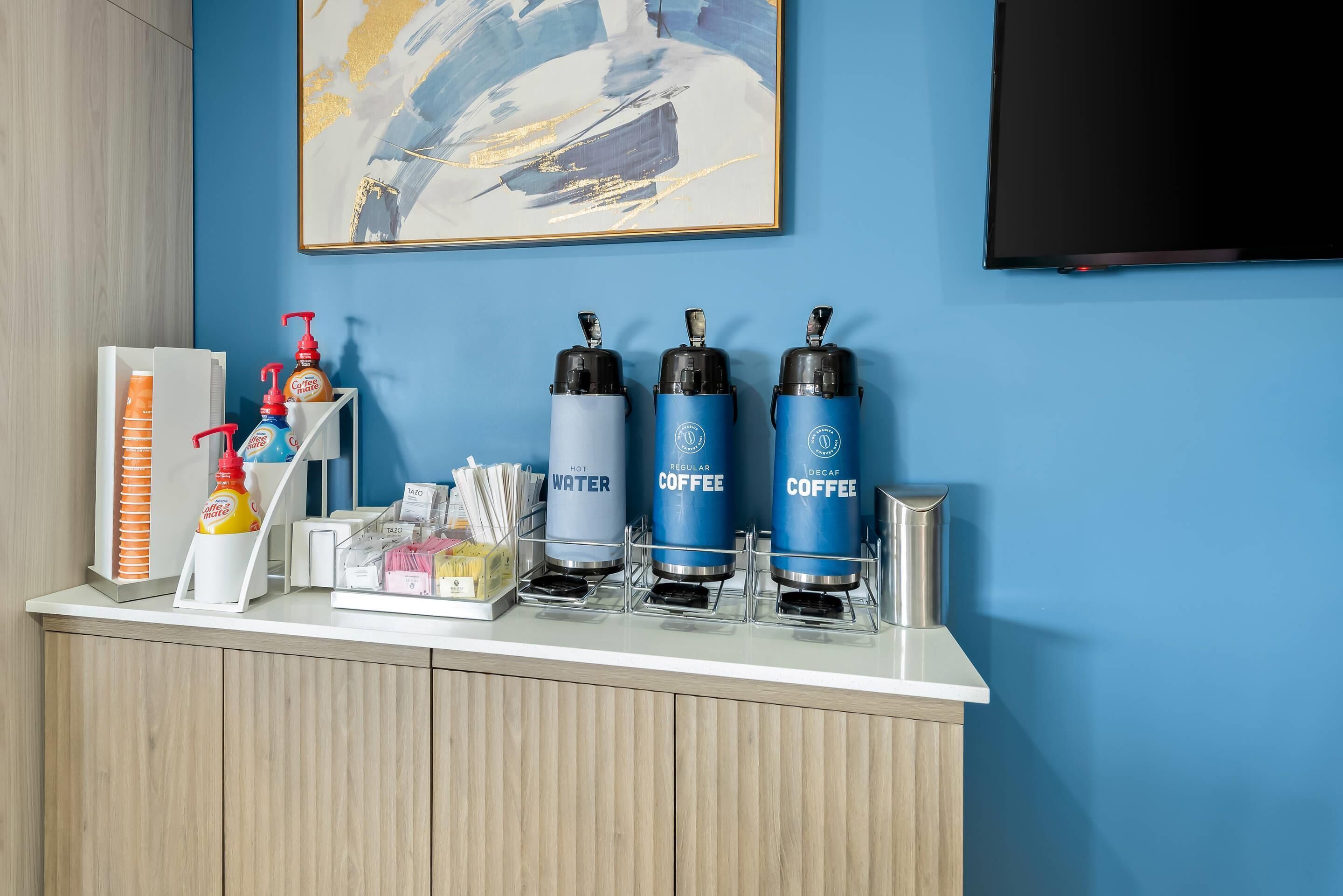 A hotel reception area with a coffee station featuring three dispensers labeled 'HOT WATER,' 'REGULAR COFFEE,' and 'DECAF COFFEE.'