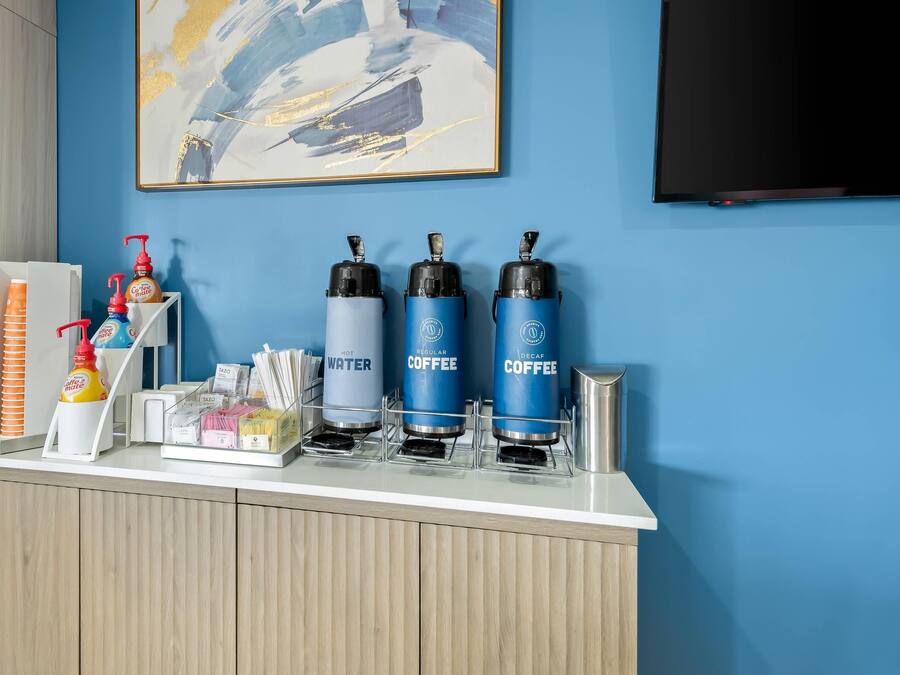 Zona d'entrada d'un hotel amb un racó de cafè que ofereix tres dispensadors etiquetats 'AIGUA CALENTA', 'CAFÈ NORMAL' i 'CAFÈ DESCAFEÏNAT'.