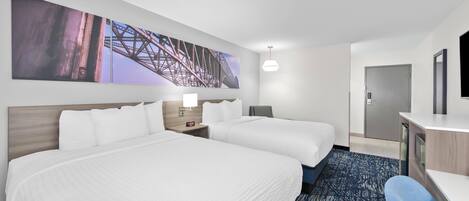 A hotel room with two beds, a desk, a chair, a large painting on the wall, and a door.