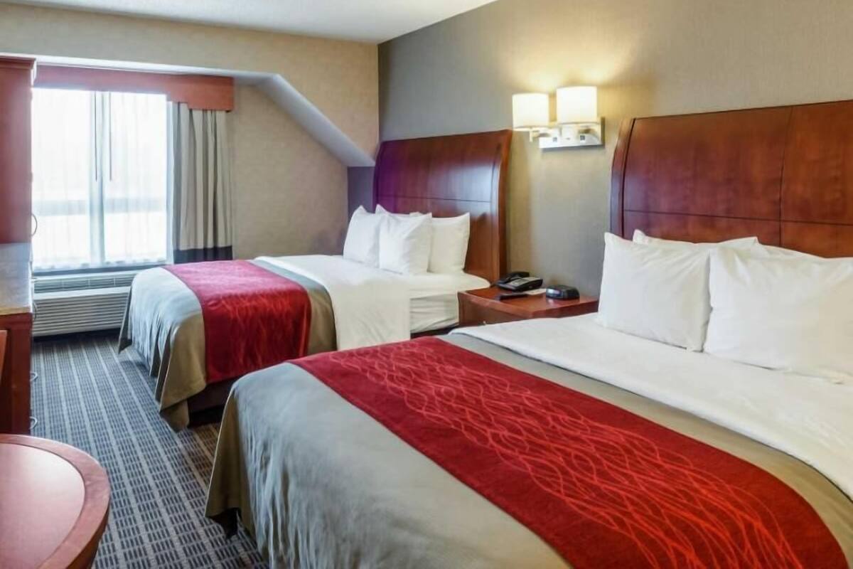 Quarto de hotel com duas camas, uma escrivaninha, uma cadeira, um abajur e uma janela com cortinas.