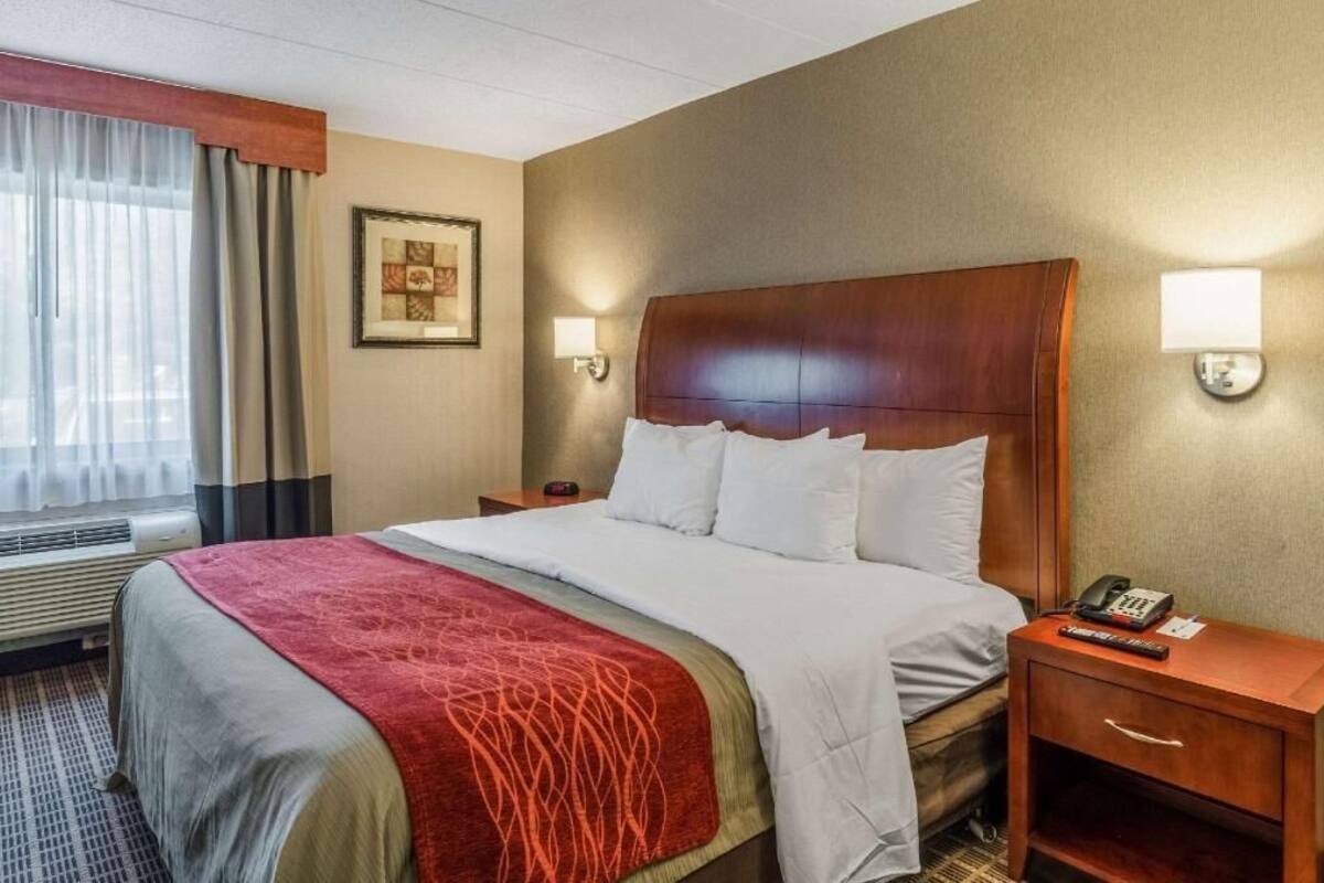 Quarto de hotel com cama, mesa de cabeceira com telefone, luminária na parede e janela com cortinas.