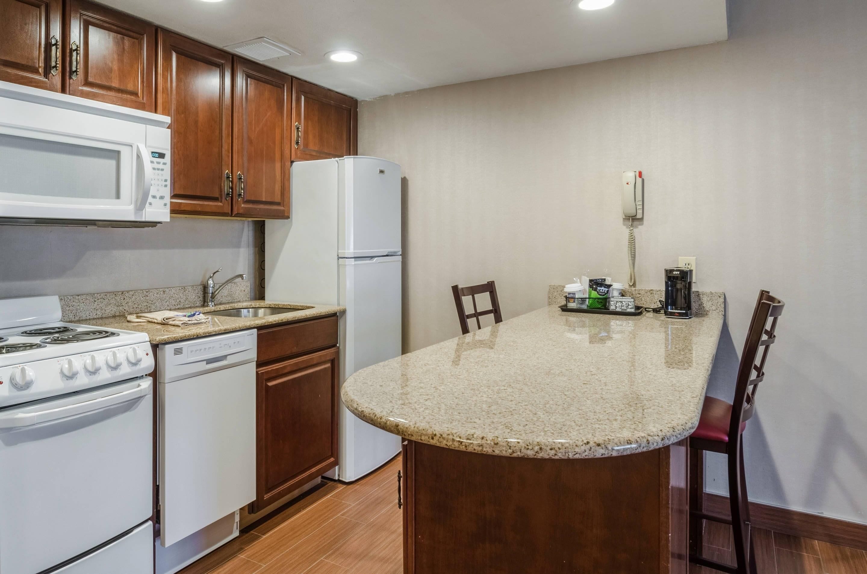 Una cocina con armarios de madera, microondas, refrigerador, estufa, lavavajillas y una isla con encimera de granito y sillas.