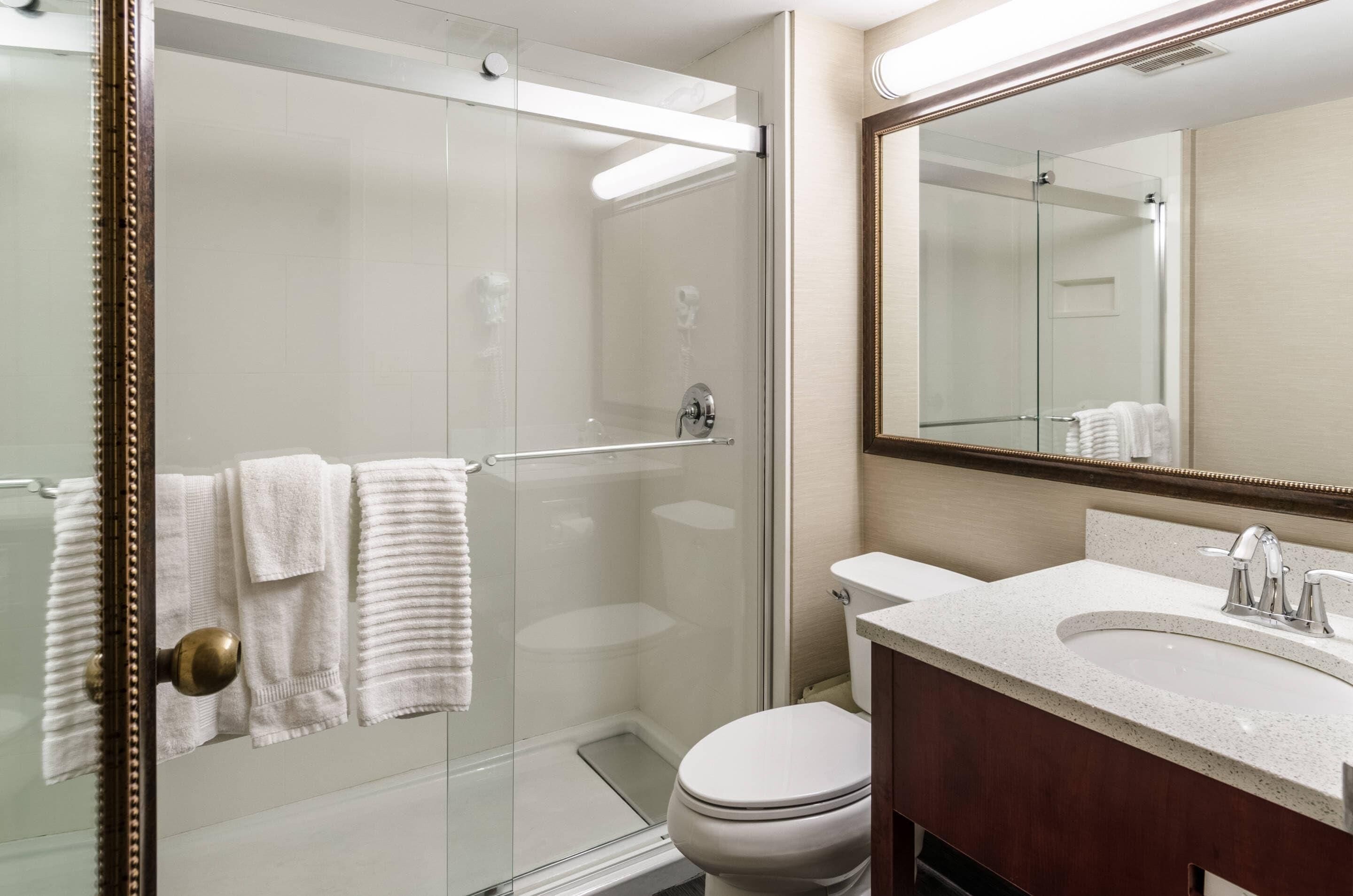 Un baño moderno con ducha de vidrio, inodoro blanco, lavamanos con espejo y toallas colgadas en un perchero.