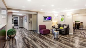 Un vestíbulo de hotel con suelo de madera, una mesa de recepción, un sillón morado, un sofá con cojines y una zona de comedor con sillas.