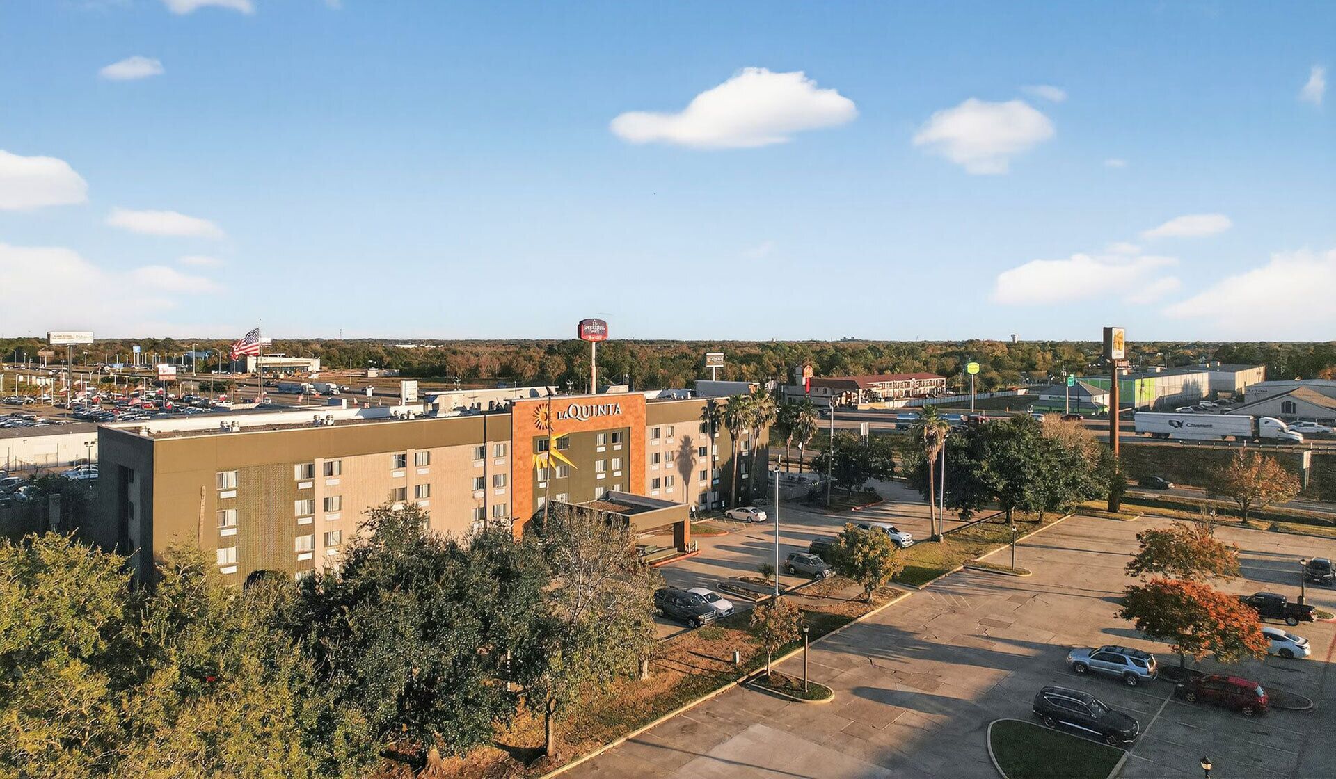 A hotel with a parking lot, surrounded by trees and a clear sky.