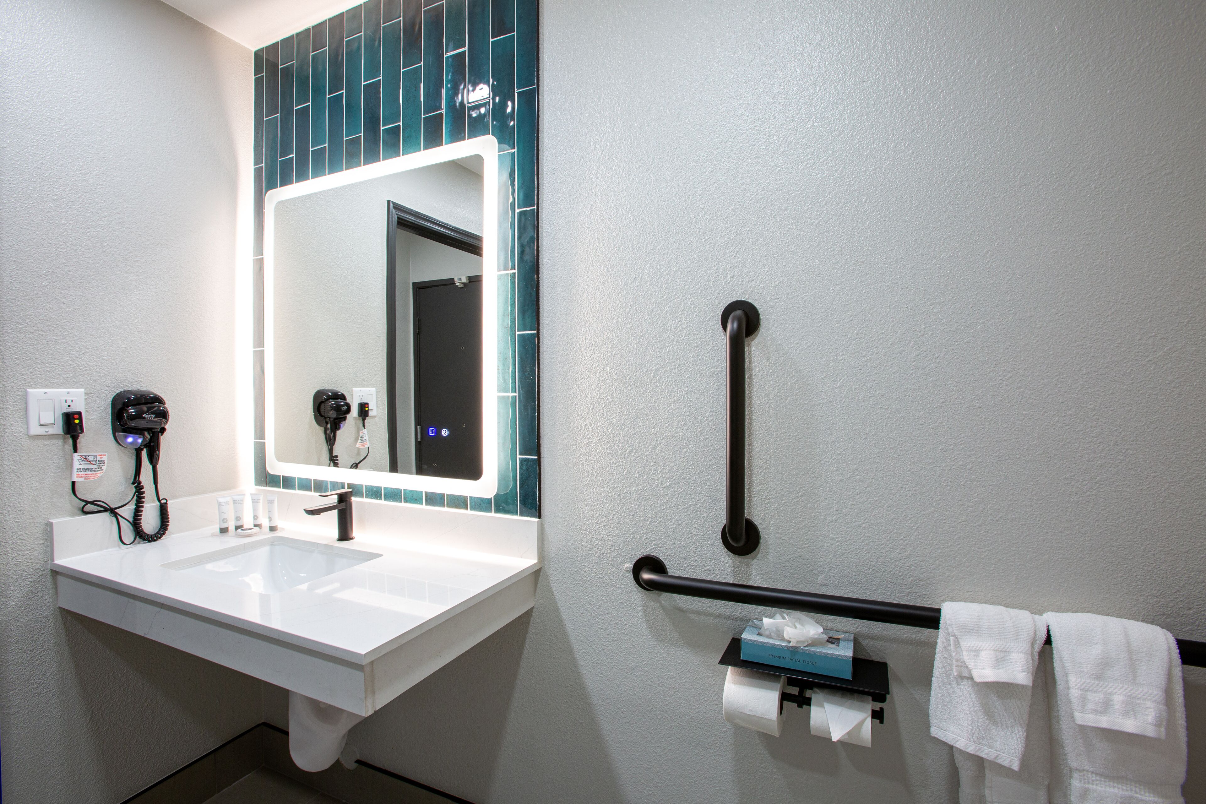 Une salle de bain avec un lavabo blanc, un miroir, un porte-serviettes et un sèche-mains.