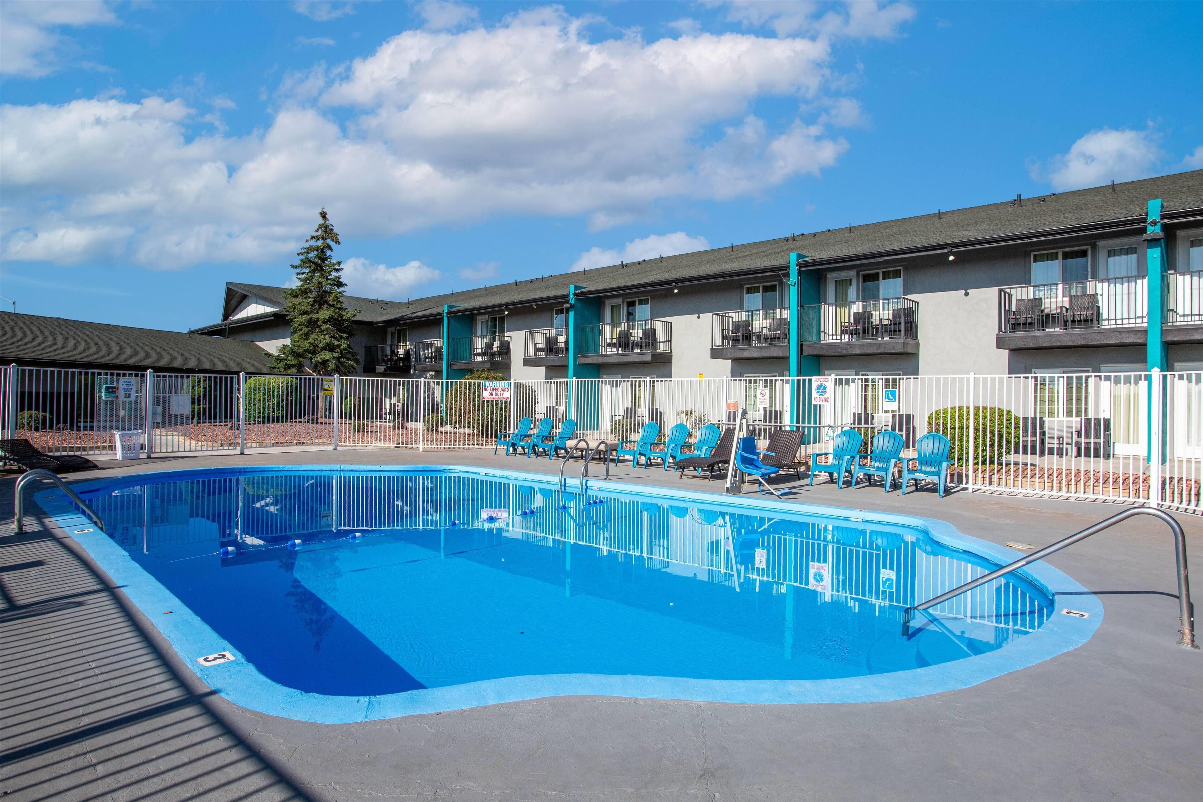 Una piscina con cerca blanca, sillas azules y un edificio con balcones al fondo.