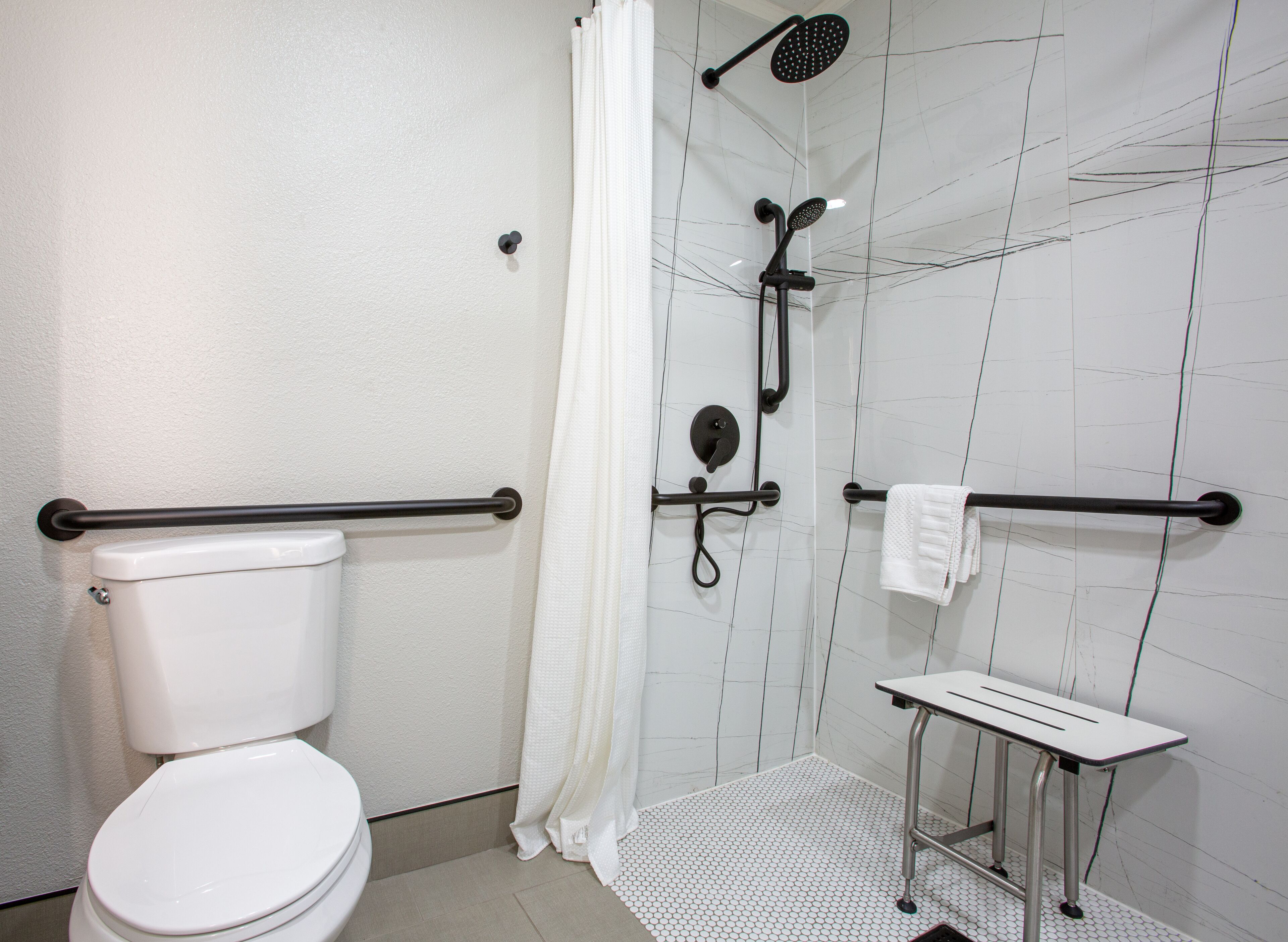 A bathroom with a toilet, a shower with a handheld showerhead, a towel rack, and a small bench.