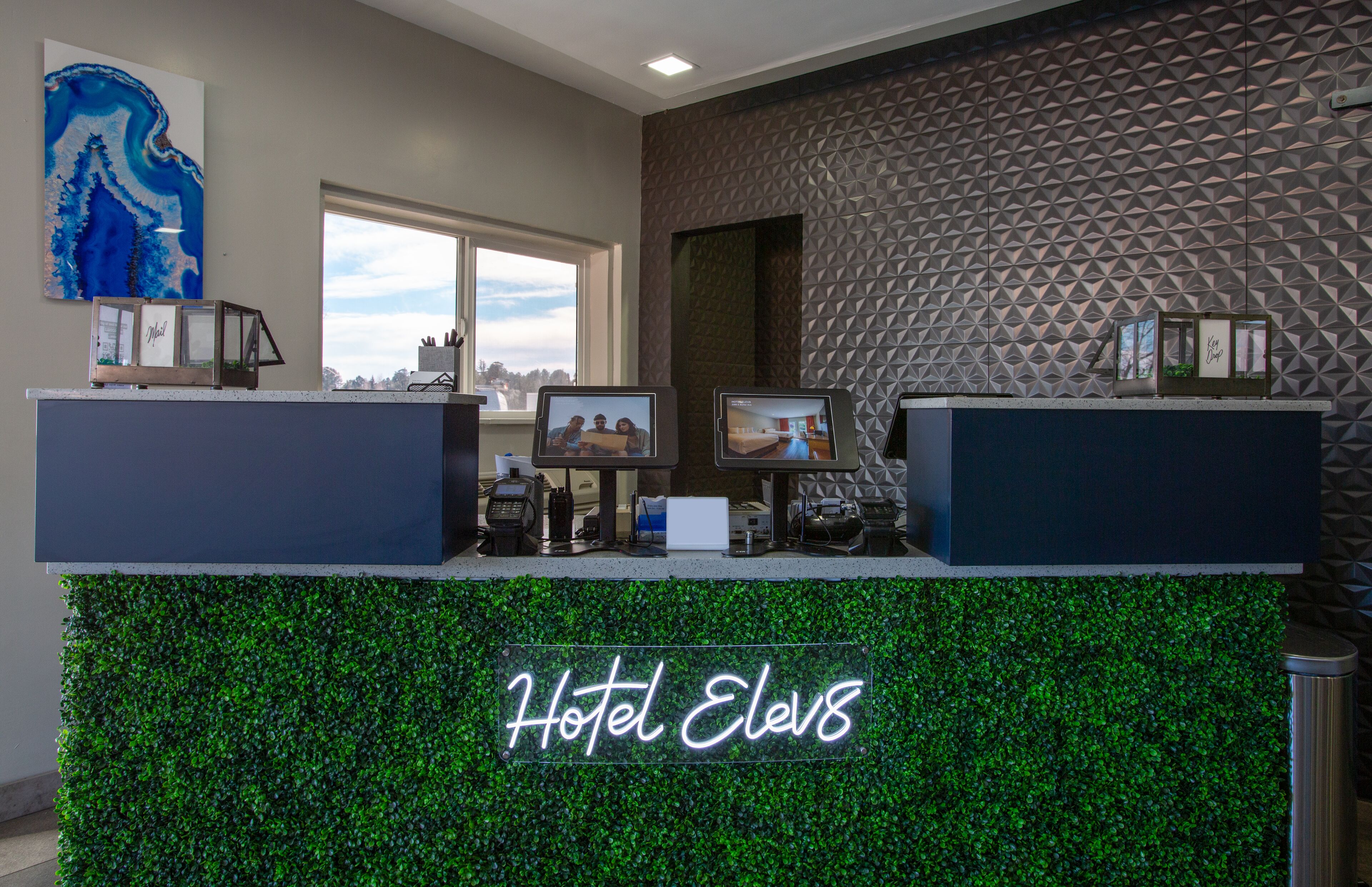 A hotel reception desk with a neon sign reading "Hotel Elev8," a large window, and a geometric patterned wall.
