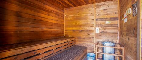 A wooden sauna with benches, a metal bucket, and a metal container on a wooden stand.