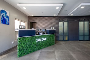 A hotel reception area with a green foliage-themed counter, a blue wall, and a geometric patterned wall in the background. - Hotel Elev8 Flagstaff, Trademark Collection by Wyndham (Flagstaff)
