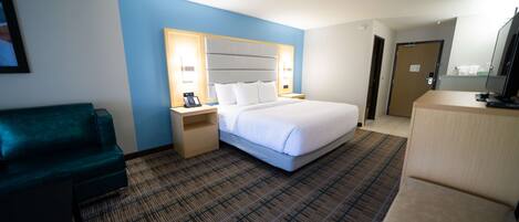 A hotel room with a large bed, a desk with a lamp, a green armchair, and a TV.