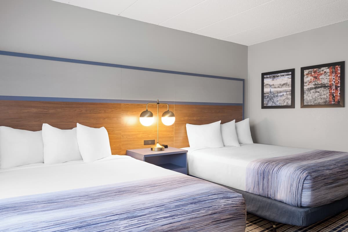 A hotel room with two beds, a wooden headboard, a nightstand with a lamp, and two framed pictures on the wall.