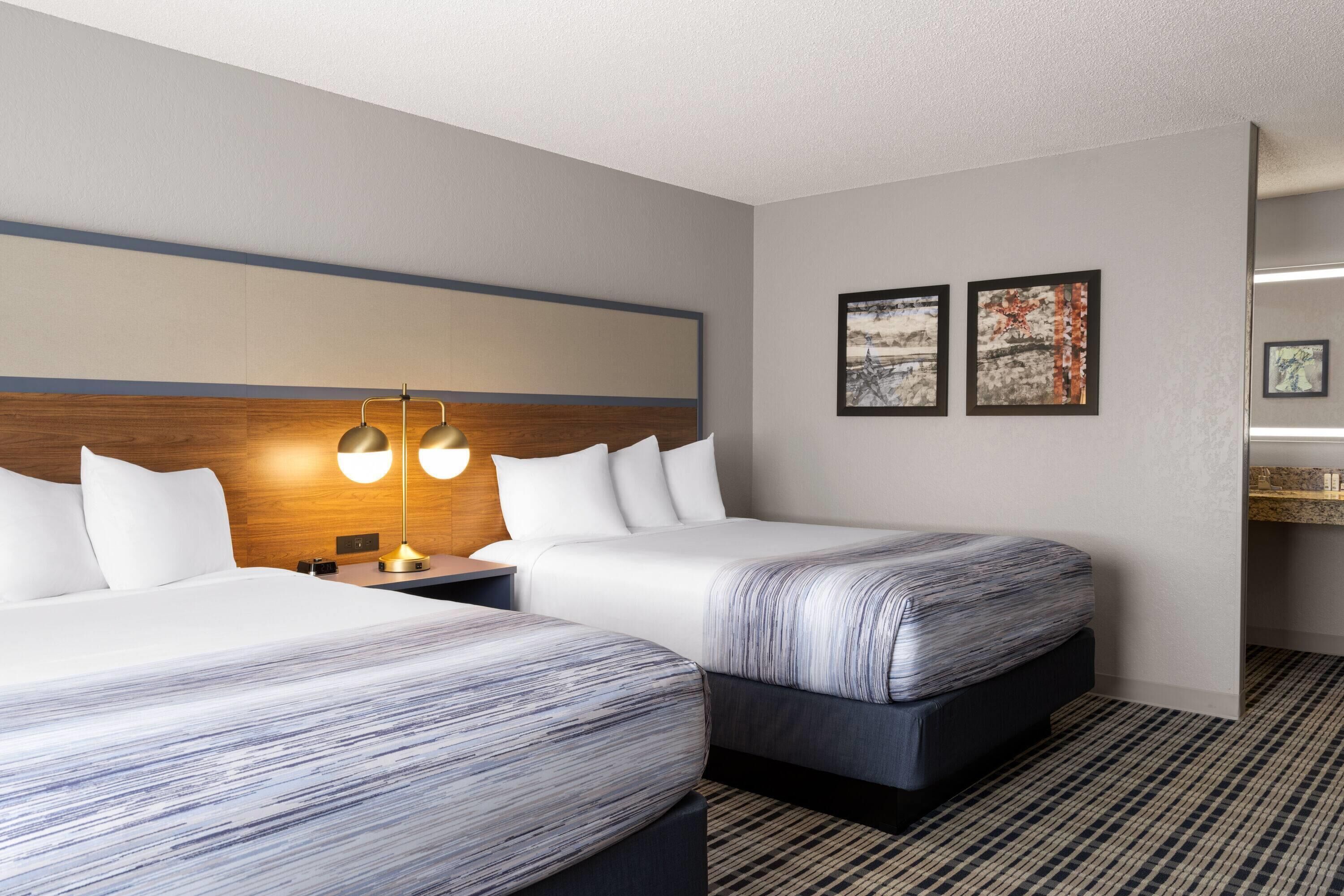 A hotel room with two beds, a wooden headboard, a nightstand with a lamp, and two framed artworks on the wall.