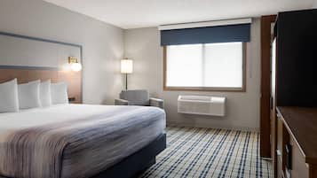 A hotel room with a large bed, a desk, a chair, a window with blinds, and a wall-mounted air conditioner.