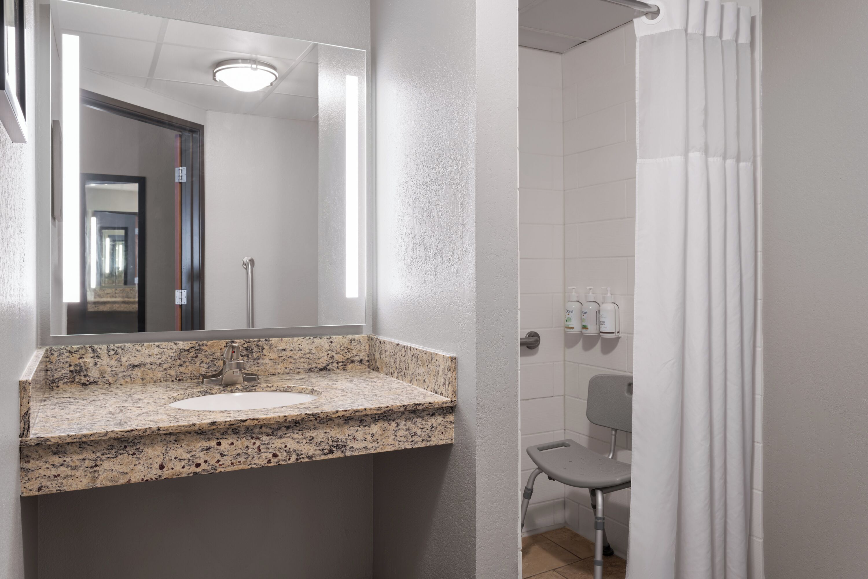 A bathroom with a granite countertop sink, a mirror, a shower curtain, and a small chair.