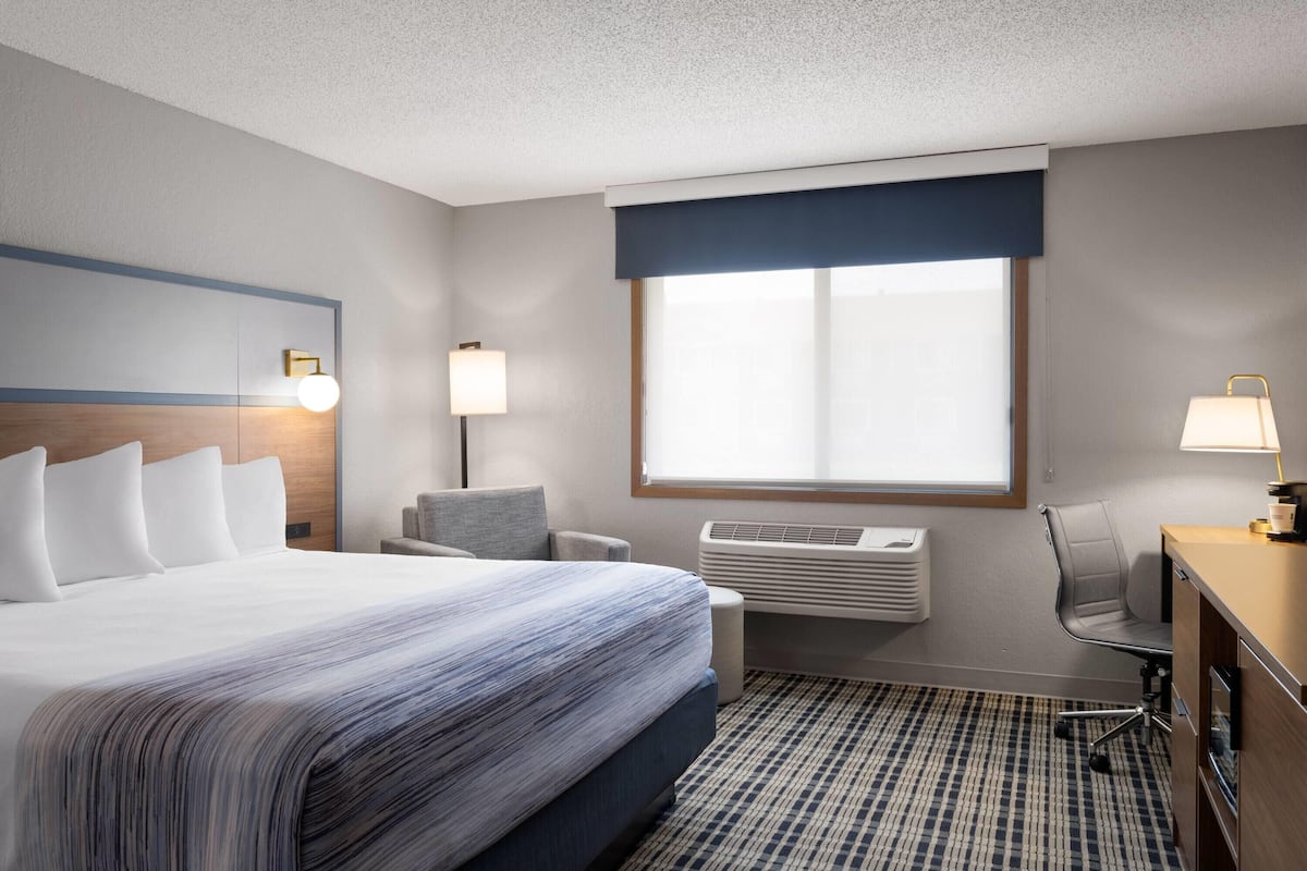 A hotel room with a large bed, a desk, a chair, a window with blinds, and a wall-mounted air conditioner.