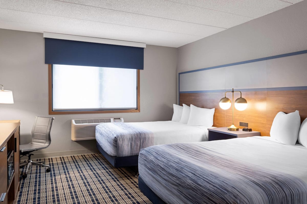 A hotel room with two beds, a desk, a chair, a large window, and a wall-mounted lamp.